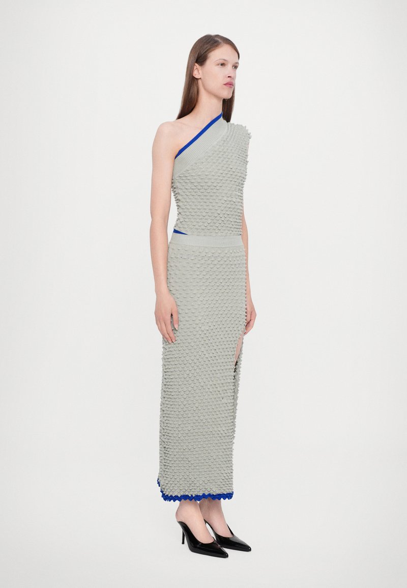 Vestido gris asimétrico con diseño texturizado, acentos azules en el escote y el dobladillo, abertura lateral, combinado con tacones altos negros.