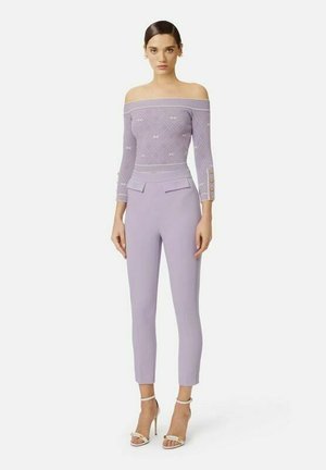 Donna in piedi indossa un top lilla con spalle scoperte e motivi geometrici, abbinato a pantaloni stretti a vita alta dello stesso colore, completati da sandali bianchi con tacco alto.