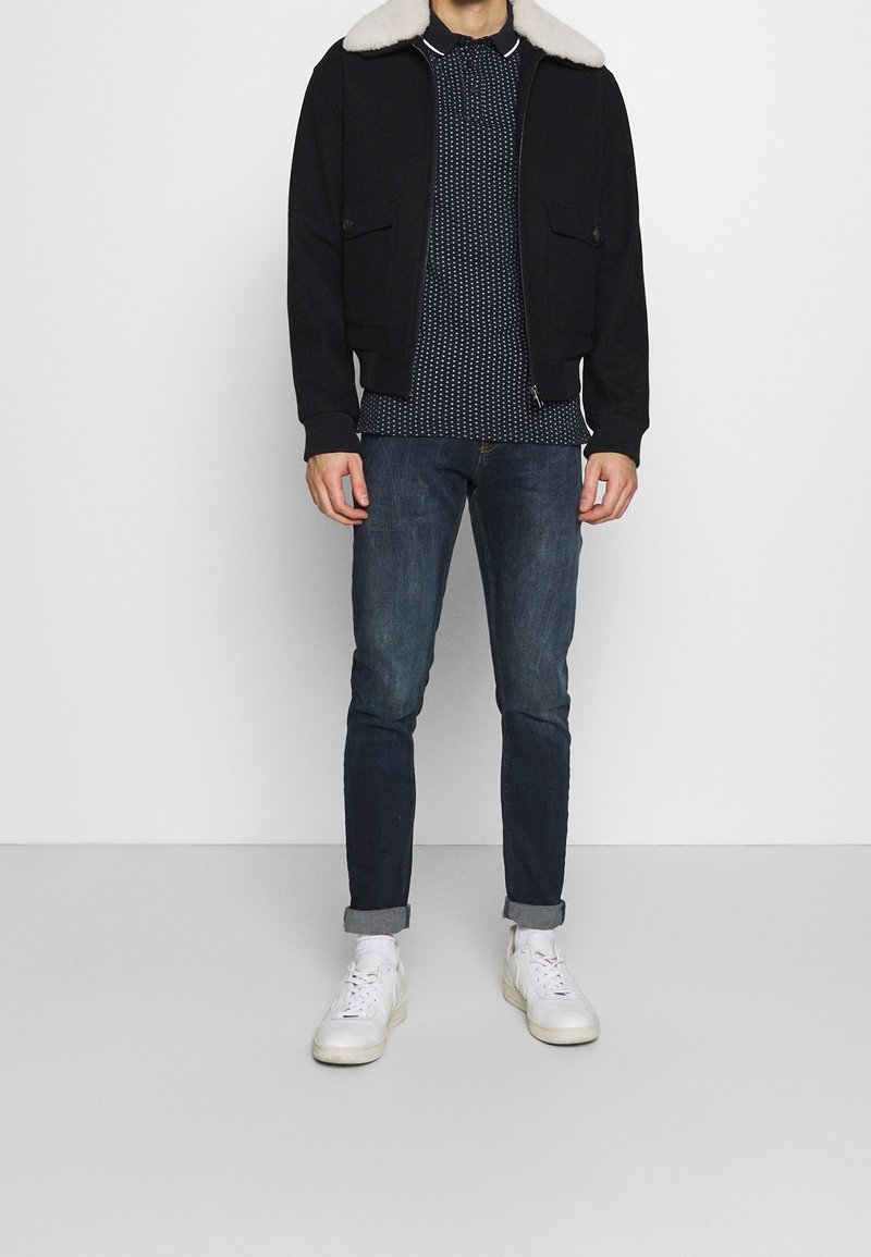 Black bomber jacket with a white shearling collar, patterned navy polo shirt, dark jeans, and white sneakers. Features a casual fit.
