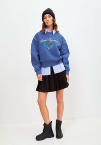 Bluza cu broderie "Bond Together" și design cu cireșe, purtată peste o cămașă în dungi light blue și alb, asortată cu o fustă plisată neagră și cizme negre.