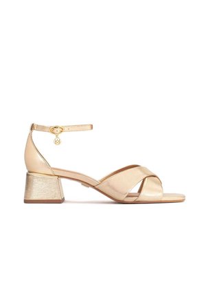 Goldfarbene metallische Sandale mit hohen Absätzen, gekreuzten Riemen, einem Knöchelriemen-Design und einem strukturierten Blockabsatz. Ein kleines Charm nahe dem Knöchel.