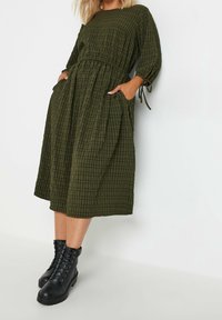 Oliivinvihreä ruudullinen midi-mekko, jossa on kireä vyötärö, lyhyet hihat, joissa on nauhat, sekä sivutaskut, yhdistettynä mustiin nauhallisiin nilkkabootseihin.