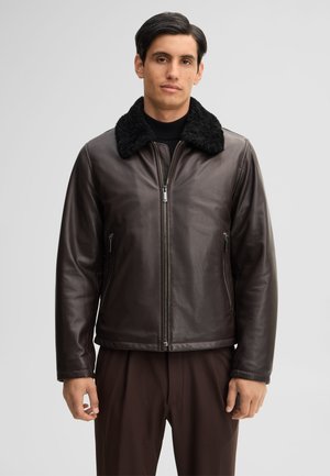 Uomo che indossa una giacca di pelle marrone scuro con colletto in pelliccia nera e pantaloni marroni, in piedi davanti a uno sfondo chiaro e uniforme.