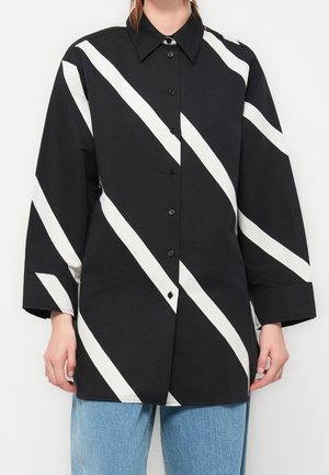 Chemise noire à manches longues boutonnée avec de larges rayures blanches diagonales portée par une personne avec un jean bleu clair.