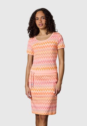 Kobieta z kręconymi włosami, ubrana w krótką sukienkę o długości do kolan z krótkim rękawem, we wzór zygzaków w kolorach różowym, pomarańczowym i żółtym, z wiązaniem w talii, na szarym tle.