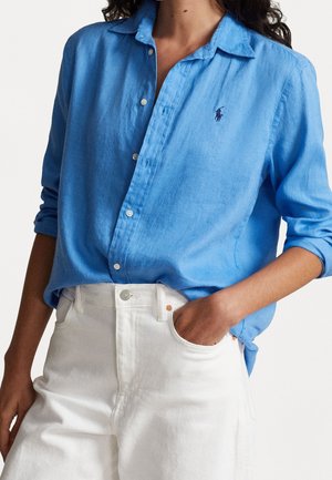 Woman wearing light blue long-sleeve button-up shirt tucked into high-waisted white pants, with one hand in pocket.