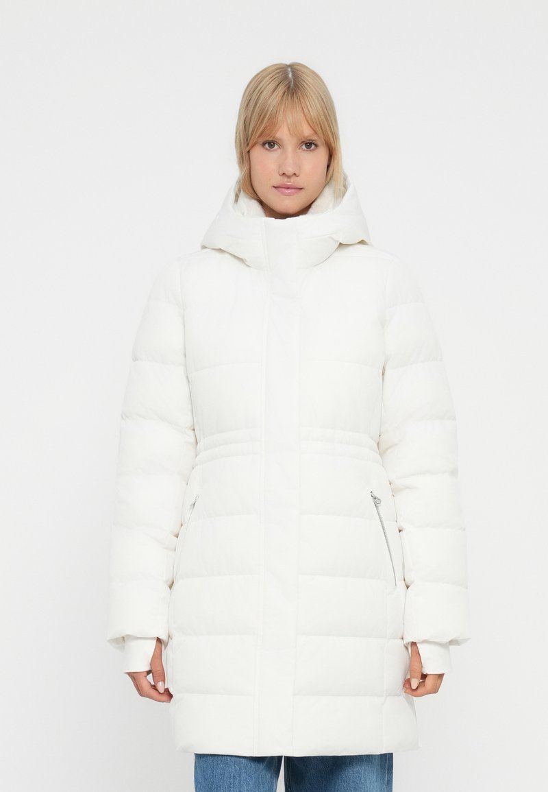 Γυναίκα που φοράει ένα άσπρο επενδυμένο παλτό με μεγάλη κουκούλα και φερμουάρ στις τσέπες, που στέκεται μπροστά σε άσπρο φόντο.