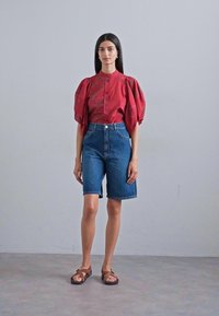 Red blouse with puff sleeves, button-up front; blue high-waisted denim shorts; brown sandals. Plain background, model standing upright.