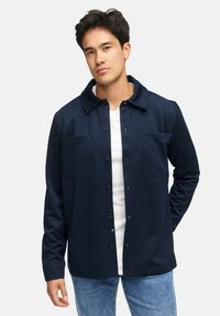 Mørkeblå button-up jakke med krave, to forlommer, lange ærmer og en afslappet pasform, båret over en hvid skjorte og blåjeans.