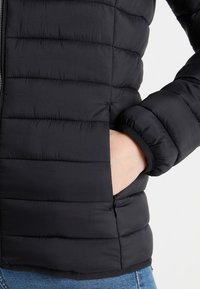 Černá péřová bunda s prošívanou texturou, se zipem vpředu a elastickými manžetami. Na levé straně je viditelná boční kapsa se zipem.