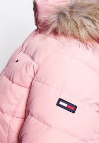Lyserød pufferjakke med struktureret overflade, hætte med pelsfor og et logo patch med røde, hvide og blå rektangulære detaljer.