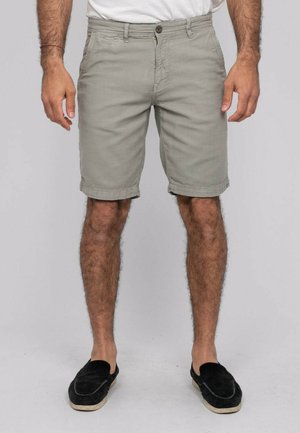 Homme portant un short gris clair arrivant aux genoux, une chemise blanche rentrée, et des chaussures noires sans lacets, debout avec les bras détendus le long du corps.