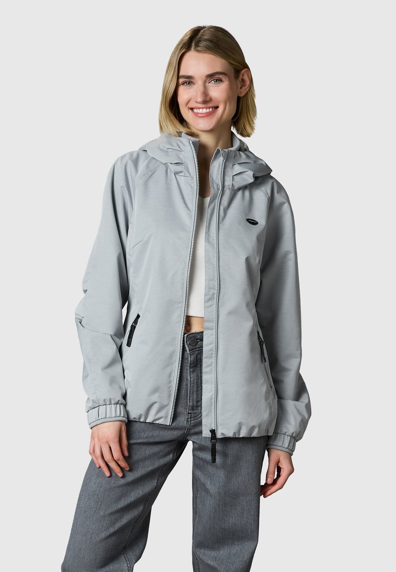 Glimlachende vrouw die een lichtgrijze capuchonjas met rits, een wit cropped topje en grijze spijkerbroek draagt, staand tegen een effen achtergrond.