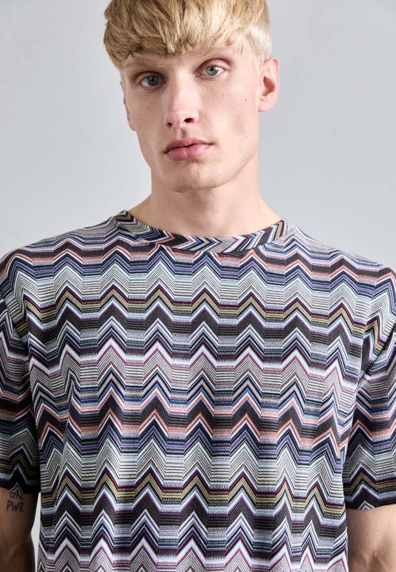Uomo biondo con occhi chiari che indossa una camicia a maniche corte con un motivo zigzag multicolore e un tatuaggio "GRL PWR" ben visibile sul braccio.