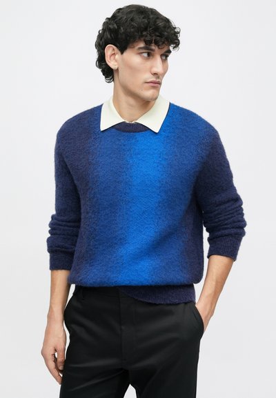 Jovem com cabelo encaracolado e escuro, vestindo um suéter felpudo azul em degradê sobre uma camisa branca com colarinho, calças pretas, com uma mão no bolso.