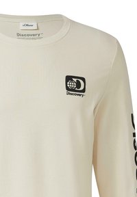 Creme lange mouw shirt van zachte stof met een zwart Discovery-logo op de linkerkant van de borst en het woord "Discovery" langs de mouw.