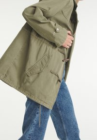 Jasnozielona kurtka wykonana z bawełny, w luźnym kroju, z zapięciem na troczki i ukośnymi kieszeniami. W połączeniu z niebieskimi dżinsami.