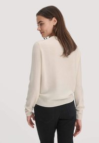 Frau mit langen braunen Haaren, die einen cremefarbenen Pullover und schwarze Hosen trägt, steht und schaut nach links vor einem schlichten Hintergrund.