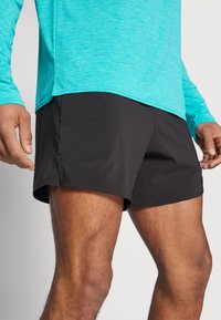Black athletic shorts with a smooth texture and a relaxed fit. Worn with a bright turquoise long-sleeve shirt.