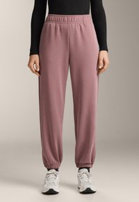 Világos rózsaszín, puha anyagból k�észült melegítőnadrág gumis derékrésszel és összehúzott bokarészsel, fehér sportcipővel és fekete hosszú ujjú felsővel párosítva.