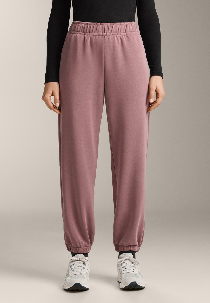 Világos rózsaszín, puha anyagból készült melegítőnadrág gumis derékrésszel és összehúzott bokarészsel, fehér sportcipővel és fekete hosszú ujjú felsővel párosítva.