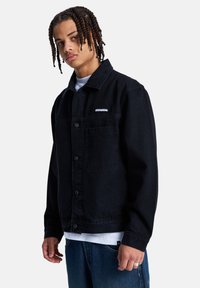 Νεαρός άνδρας με πλεξούδες, που φοράει μαύρο σακάκι με κουμπιά πάνω από λευκό πουκάμισο και μπλε τζιν, στέκεται μπροστά σε απλό φόντο.