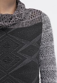 Pull noir et gris avec un corps en tricot texturé, un col cheminée à motifs et des textures de tissu variées sur les épaules et les manches.
