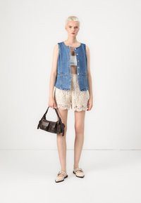 Denim ermeløs jakke med knapper foran, kombinert med beige blonde shortser og en mørk brun skulderveske. Lyse sko fullfører antrekket.
