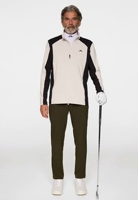 Completo da golf maschile con giacca con zip di colore beige chiaro e nero, pantaloni verdi e scarpe da golf bianche, con in mano una mazza da golf.