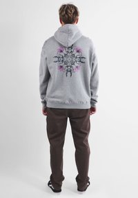 Felpa con cappuccio grigia con un design stampato colorato sul retro, abbinata a pantaloni marroni scuri e sneaker nere.