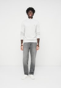 Grå genser med høy hals, ribbestrikkede mansjetter og liten logo, kombinert med grå jeans og hvite joggesko, stående mot en ensfarget bakgrunn.