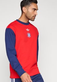 Κόκκινο και ναυτικό φούτερ με μακριά μανίκια, ριμπ μανσέτες και στρογγυλή λαιμόκοψη. Διαθέτει το λογότυπο της Adidas και το έμβλημα της FC Bayern Munich στο στήθος.