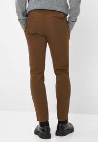 Pantaloni slim fit marroni realizzati in tessuto liscio, dotati di tasche posteriori e cintura, abbinati a scarpe nere e calze scure.