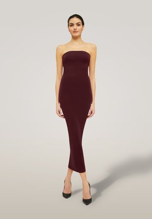 Vrouw staat in een strapless, aansluitende midi-jurk in donker bordeaux met zwarte hakken tegen een eenvoudige lichte achtergrond.