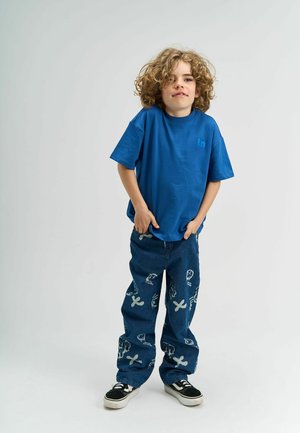 Niebieska bawełniana koszulka o luźnym kroju w zestawieniu z dżinsami o luźnym kroju z białymi nadrukami graficznymi. Czarne sneakersy dopełniają stylizację.