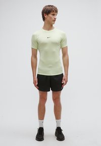 Ανοιχτό πράσινο αθλητικό μπλουζάκι με κοντά μανίκια, συνδυασμένο με μαύρο σορτς. Το μοντέλο φοράει μαύρα αθλητικά παπούτσια και λευκές κάλτσες πλήρους μήκους. Μινιμαλιστικός σχεδιασμός.