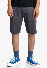 Pantaloni corti grigi in cotone con taglio dritto, tasche laterali e orli rifiniti. Indossati con scarpe da ginnastica blu e calze blu.