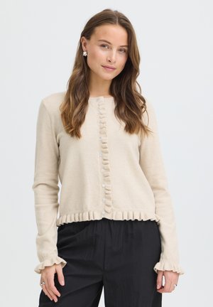 Frau mit langen, welligen Haaren, die eine beige, mit Knöpfen versehene Strickjacke mit Rüschen an den Kanten und schwarze Hosen trägt, steht vor einem schlichten weißen Hintergrund.