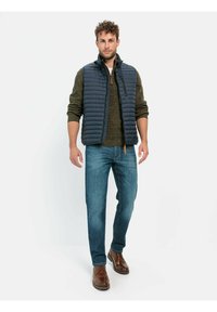 Marineblaue Steppweste mit hohem Kragen über einem grünen Strickpullover, kombiniert mit blauen Jeans und braunen Lederschuhen. Dünne, gestepte Textur.
