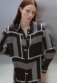 Camicia marrone con bottoni e motivi lineari bianchi. Presenta un colletto aperto e maniche lunghe, realizzata in tessuto morbido con una vestibilità comoda.