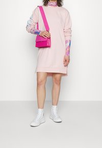 Světle růžové šaty s dlouhými rukávy, zdobené barevnými vzory na rukávech. Spárované s růžovou crossbody taškou a bílými vysokými teniskami.
