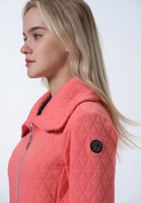 Koraal gewatteerde jas met een brede kraag, voorzien van een ritssluiting en een ronde zwarte logo patch op de mouw. Soepele textuur, ruitpatroon.