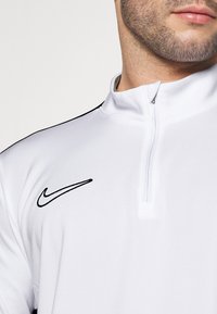 Cămașă albă cu fermoar, realizată dintr-un material neted, având un logo Nike negru pe pieptul stâng și accente negre pe umeri.