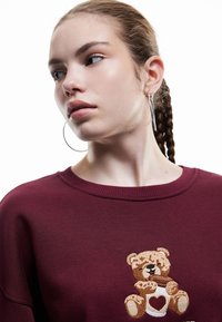 Sudadera burdeos con un oso de peluche bordado que sostiene una taza en forma de corazón. Tela suave, cuello redondo y corte relajado.