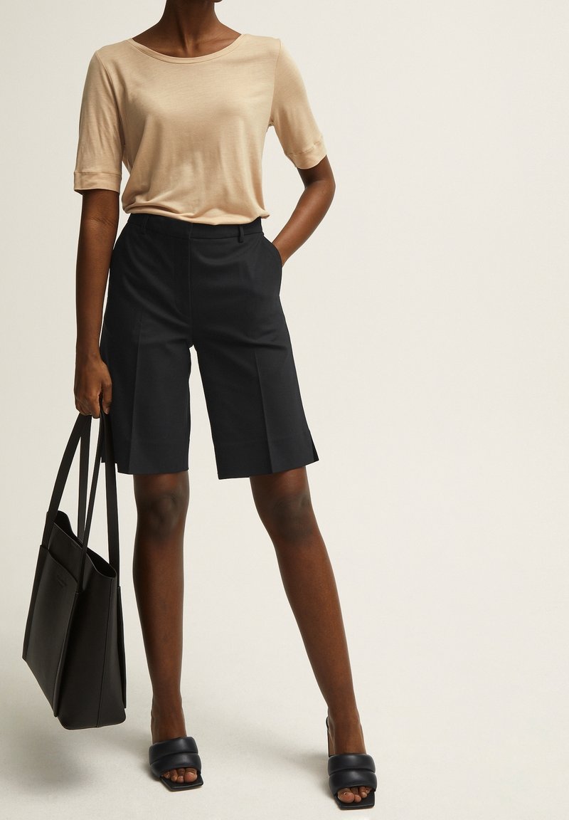 Haut beige à manches courtes avec un col rond, associé à un short ajusté de couleur foncée avec des poches latérales. Le modèle tient un sac fourre-tout noir et porte des sandales noires.