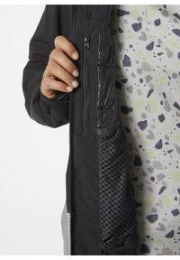 Schwarze Jacke mit Reißverschlusstaschen, strukturiertem Stoff und Mesh-Futter. Das Innenfutter weist ein gesprenkeltes Muster in Grau, Grün und Marineblau auf.