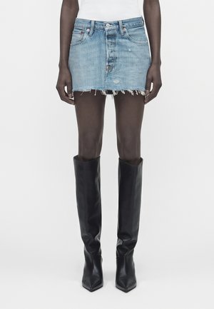 Personne aux jambes fines portant une mini-jupe en denim bleu clair usée, associée à des bottes en cuir noires montantes jusqu'aux genoux, debout devant un fond uni.