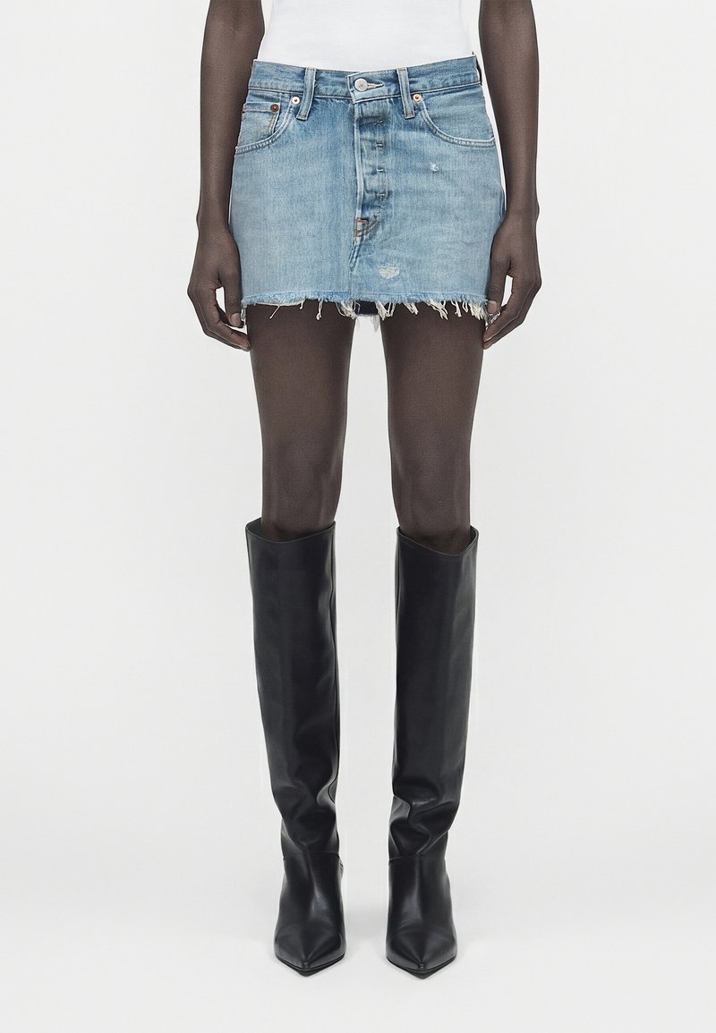 Άτομο με λεπτά πόδια φοράει μια φθαρμένη ανοιχτό μπλε μίνι φούστα από τζιν, συνδυασμένη με μαύρες δερμάτινες μπότες μέχρι το γόνατο, στέκεται σε απλό φόντο.