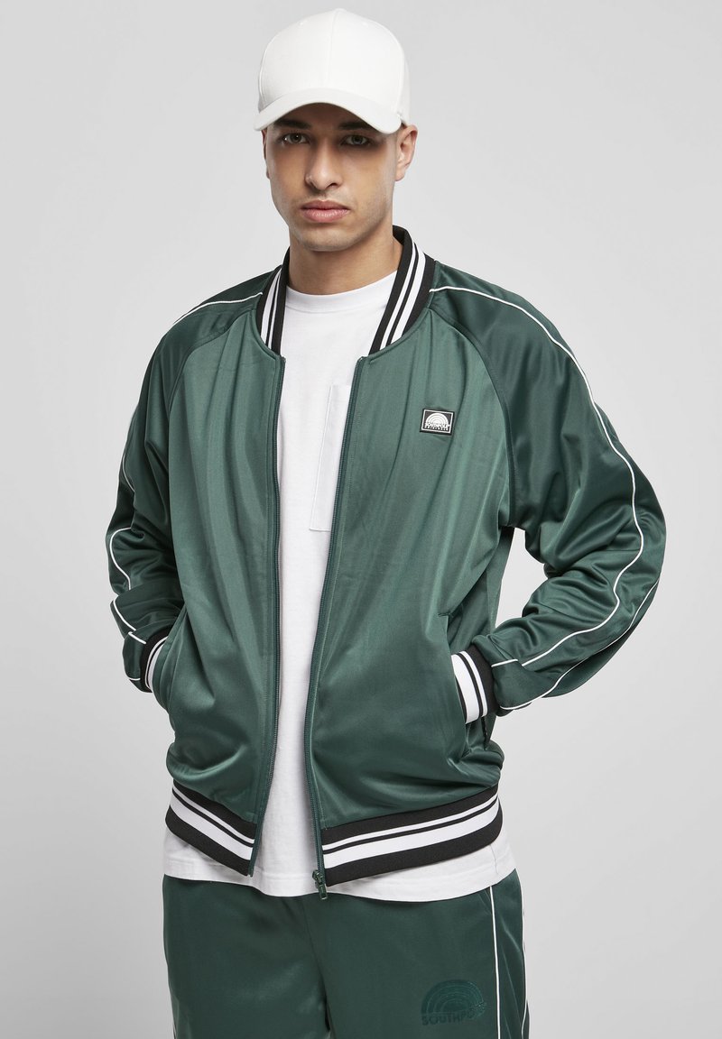 Giacca bomber in satin verde con dettagli a strisce bianche, polsini e orlo nero a costine. Abbinata a una t-shirt bianca e pantaloni coordinati.