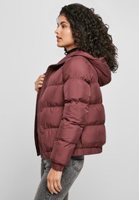 Donna con capelli ricci che indossa un giubbotto imbottito borgogna e jeans grigi, rivolta a sinistra, mentre tiene la zip del giubbotto vicino al petto.